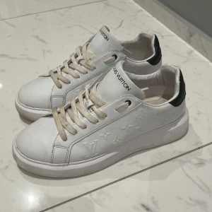 Vita sneakers från Louis Vuitton - Snygga vita sneakers från Louis Vuitton med klassisk design och diskret logotypprägling på sidan. Skorna har snörning och en svart detalj vid hälen. Perfekta för en stilren look.