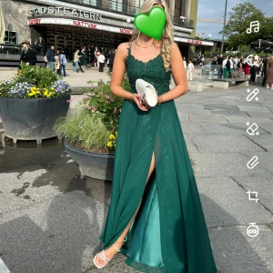 Grön långklänning med spetsdetaljer - Säljer nu min älskade balklänning, använd en gång. Elegant grön långklänning med spetsdetaljer och smala axelband. Klänningen har en slits framtill och en öppen rygg med snörning. Perfekt för er som ska på bal eller liknadade! 