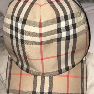 Rutig keps från Burberry - Snygg rutig keps från Burberry i klassiskt beige, svart och rött mönster. Perfekt för att ge din outfit en stilren touch. Kepsen har en normal passform 