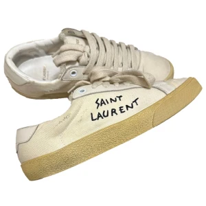 Beige sneakers från Saint Laurent - Snygga beige sneakers från Saint Laurent med handskriven logga på sidan. Skorna har en klassisk design med snörning och en robust gummisula. Perfekta för en stilren och avslappnad look.