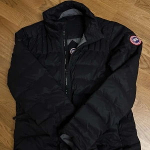 Svart dunjacka från Canada Goose - Säljer en svart dunjacka från Canada Goose med dragkedja och långärmad design. Jackan har en klassisk look med varumärkets logga på ärmen. Perfekt för kalla dagar. Ett litet hål som inte syns så mycket