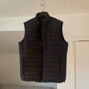 Svart dunväst från Peak Performance - Stilren svart dunväst från Peak Performance. Västen har en dragkedja framtill och är quiltad för extra värme. Perfekt för lager-på-lager under kyliga dagar. Den har en klassisk passform och är lätt att matcha med olika outfits.