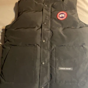 Svart dunväst från Canada Goose - Säljer en svart dunväst från Canada Goose med deras ikoniska märke på bröstet. Västen har knappar framtill och två stora fickor. Perfekt för kyliga dagar när du vill hålla värmen utan att kompromissa med stilen.