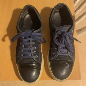 Marin blå sneakers från Lanvin - Snygga svarta sneakers från Lanvin med blå snörning och vit sula. Skorna har en stilren design med en glansig tådel och flera snörhål för justerbar passform.