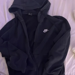 Svart hoodie från Nike - Säljer en svart hoodie från Nike med dragkedja och huva. Den har en klassisk design med Nike-loggan på bröstet. Perfekt för en avslappnad stil, mer som en kofta! I helt nytt skick. Köpte för en vecka sen aldrig använd!