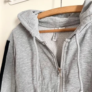 Tjocktröja - Ljusgrå tjocktröja med huva och svarta linjer längst ärmarna! Tröjan har använts fåtal gånger, och är i väldigt bra skick. Sitter som M. Alla använda plagg tvättas noga innan leverans! Det är bara att skriva om du har några frågor! <3 
