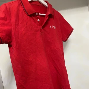 Röd pikétröja från Armani - Snygg röd pikétröja från Armani med korta ärmar och klassisk krage. Tröjan har knappar framtill och en liten logga på bröstet. Perfekt för en avslappnad stil.