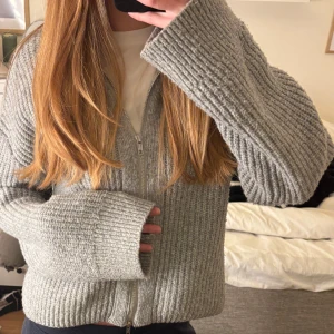 Grå stickad kofta med dragkedja - Säljer en mysig grå stickad kofta med dragkedja framtill. Koftan har långa ärmar och en ribbad struktur som ger en stilren look. Perfekt för kyliga dagar!