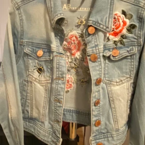 Cool jeansjacka från LAB Industries - Supersnygg jeansjacka från LAB Industries med broderade blommor och bin. Jackan har en ljusblå tvätt och knappar framtill. Perfekt för att ge din outfit en cool och unik touch! Säljes i fint skick!