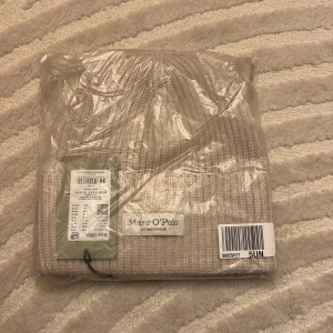Beige stickad mössa från Marc O'Polo - Säljer en stilren beige stickad mössa från Marc O'Polo. Mössan har en ribbad design och en diskret etikett framtill. Perfekt för att hålla värmen under kyliga dagar.
