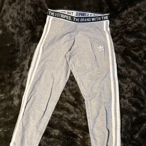 Grå tights från Adidas - Snygga grå tights från Adidas med klassiska vita ränder längs sidorna. Byxorna har en elastisk midja med texten 'THE BRAND WITH THE 3 STRIPES'.