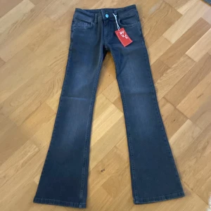 Grå bootcut jeans från RYVLS - Snygga grå bootcut jeans från RYVLS med medel midja och dubbla knappar fram. Jeansen har fickor med knappar bak och en klassisk femficksdesign. Perfekta för en stilren look. Storlek 146 (barn)