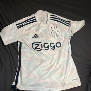 Vit Ajax fotbollströja från Adidas - Säljer en vit Ajax fotbollströja från Adidas med korta ärmar. Tröjan har ett subtilt mönster och Ajax-loggan med tre stjärnor på bröstet. Den har även mörkblå detaljer och Ziggo som sponsortryck på framsidan. Jätte bra skick och knappt använd. Modellen är 170cm cirka.