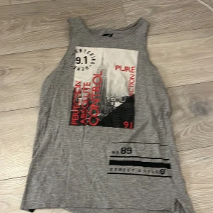 Grå ärmlös topp med tryck - Säljer en grå ärmlös topp med tryck i svart och rött. Toppen har en urban stil med text och grafiska element på framsidan. Perfekt för en avslappnad look.