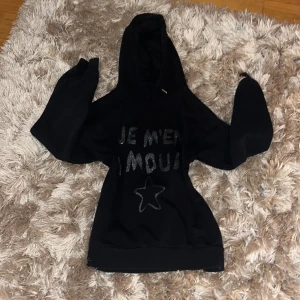 Svart hoodie från Gina Tricot - Säljer en svart hoodie från Gina Tricot med texten 'Je m'en Amour' och en stjärna i glittrigt tryck på framsidan. Perfekt för en avslappnad stil med en mysig huva och långa ärmar. Passar bra till både jeans och leggings. Den är i storlek 130/146 men passar även för 152. Jag säljer den eftersom att den är lite för liten på mig😔har använt ett par fåtal gånger men den är i bra kvalitet!! Köpte för 300kr men säljer för ungefär 150kk💞Priset kan diskuteras🥂💗