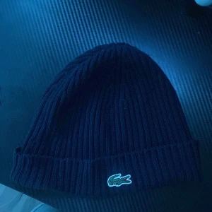 Mörkblå ribbad mössa från Lacoste - Snygg mörkblå ribbad mössa från Lacoste med det klassiska krokodilmärket. Tillverkad i mjukt material för extra komfort.