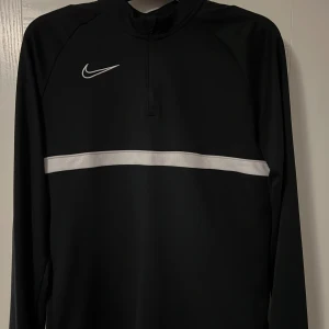 Svart långärmad tröja från Nike - Säljer en stilren svart långärmad tröja från Nike med en vit rand över bröstet och en liten dragkedja vid halsen. Perfekt för träning eller vardagsbruk. Nike-loggan är snyggt placerad på bröstet. Passar bra till både jeans och träningsbyxor.