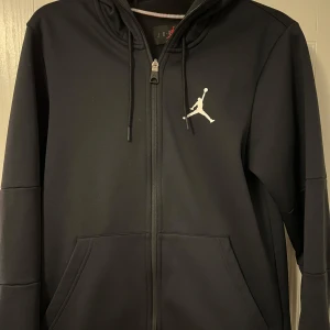 Svart hoodie från Jordan - Säljer en stilren svart hoodie från Jordan med dragkedja och justerbar huva. Den har en vit logotyp på bröstet och långa ärmar. Perfekt för en sportig look eller avslappnade dagar.