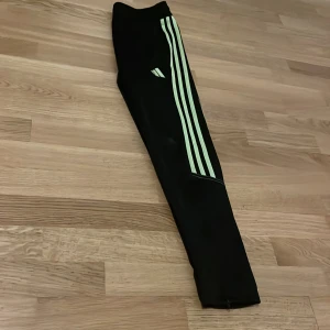 Svarta träningsbyxor från Adidas - Snygga svarta träningsbyxor från Adidas med klassiska gröna ränder längs sidorna. Perfekta för träning eller en avslappnad dag. Byxorna har en bekväm passform och är tillverkade i ett mjukt material.