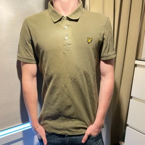 Olivgrön pikétröja från Lyle & Scott - Säljer en stilren olivgrön pikétröja från Lyle & Scott med klassisk krage och tre knappar. Tröjan har en broderad logotyp på bröstet och korta ärmar. Perfekt för en avslappnad look.