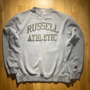 Grå sweatshirt från Russell Athletic - Säljer en klassisk grå sweatshirt från Russell Athletic med tryck på framsidan. Tröjan har långa ärmar och ribbade muddar vid ärmslut och nederkant. Perfekt för en avslappnad stil.🤍Vintage🌟