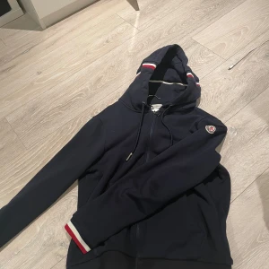 Mörkblå huvtröja från Moncler - Säljer en snygg mörkblå huvtröja från Moncler med dragkedja och ribbade muddar. Tröjan har en broderad logotyp på bröstet och detaljer i rött och vitt på ärmslut och huva. Perfekt för en stilren look.