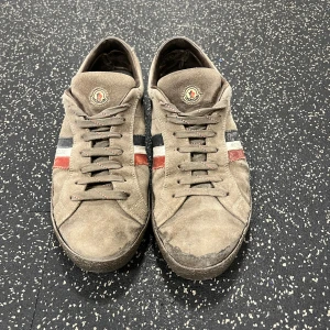 Bruna sneakers från Moncler - Säljer ett par bruna sneakers från Moncler i mocka med snörning. Skorna har Monclers logotyp på plösen och ränder i rött, vitt och blått på sidorna. Perfekta för en stilren look.