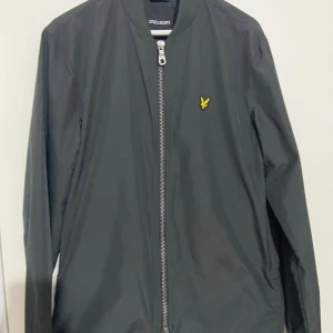 Grå/svart bomberjacka från Lyle & Scott - Snygg svart/grå bomberjacka från Lyle & Scott med dragkedja och ribbad krage. Jackan har ett broderat gult emblem på bröstet och är perfekt för en stilren look. Passar bra till både vardag och mer avslappnade tillfällen. Passar både för S och M. Ordpris: 1300kr. Går och diskutera om priset 