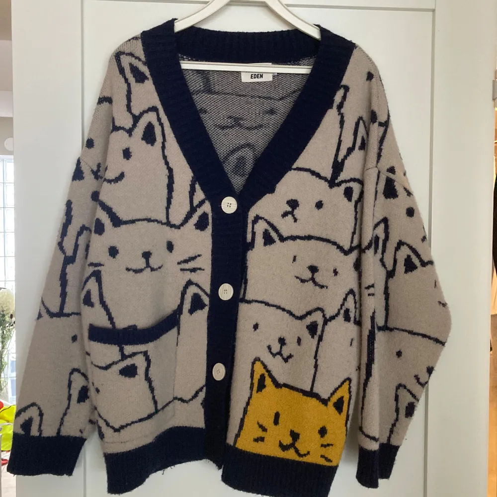 Säljer en unik beige kofta från Aelfric Eden med ett charmigt kattmönster i marinblått och en gul kattdetalj. Koftan har långa ärmar och stora knappar framtill. Något oversized. . Neuletakit & Villapaidat.