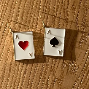 Örhängen med spelkort - örhängen formade som spelkort med essmotiv. Ena örhänget har ett rött hjärta och det andra ett svart spader, båda på vit bakgrund. Är lite tunga samt stora