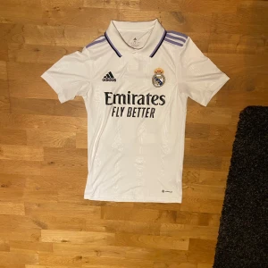 Vit Real Madrid fotbollströja från Adidas - Säljer en vit Real Madrid fotbollströja från Adidas med korta ärmar. Tröjan har klubbens emblem och sponsorlogga på framsidan samt spelarnamnet 'VINI JR.' och nummer 20 på baksidan. Perfekt för fans av laget!