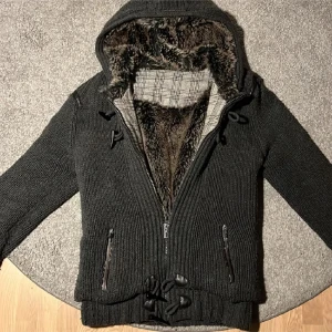 Fodrad cardigan - Säljer en svart stickad jacka med mysigt pälsfoder och rutigt innerfoder. Jackan har dragkedja och knappar framtill samt ribbade muddar. Väldigt skön, bra skick. Säljer pga fel storlek