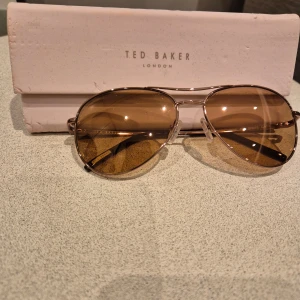 Solglasögon från Ted Baker - Snygga solglasögon från Ted Baker med en klassisk pilotform. De har en tunn metallram i guld och bruna tonade glas. Perfekta för en stilren look.