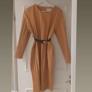 Beige klänning från Creative Collective - Snygg beige klänning från Creative Collective med långa ärmar och ett stilrent bälte i midjan. Klänningen har en dragkedja i ryggen och plisserade detaljer på ärmarna och nederdelen. Perfekt för en chic look!Bälte ingår inte I klänningen. 