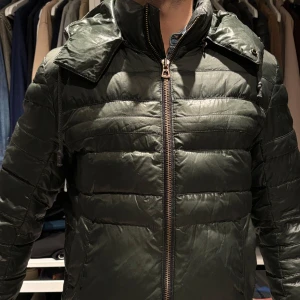 Hugo Boss Grön quiltad pufferjacka. 48 - Säljer en grön quiltad pufferjacka med dragkedja och avtagbarhuva. Jackan har en stilren design med långa ärmar och är perfekt för kyligare väder. Den är både praktisk och snygg, idealisk för att hålla värmen. M/48
