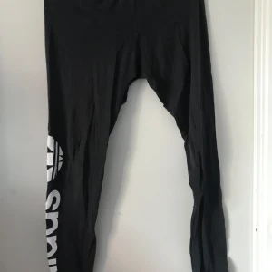 Svarta leggings från Adidas - Snygga svarta leggings från Adidas med stort vitt logotryck längs benet. Perfekta för träning eller en avslappnad dag. De är stretchiga och bekväma, vilket gör dem till ett bra val för olika aktiviteter.