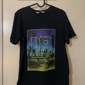Svart t-shirt med LA-tryck från H&M - Cool svart t-shirt från H&M med ett färgglatt LA-tryck på framsidan. Perfekt för en avslappnad stil med palmer och stadssilhuett i bakgrunden. Gjord i mjukt material för bekväm passform.