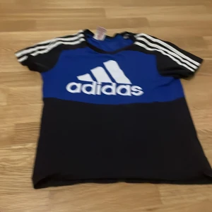Blå och svart t-shirt från Adidas - Säljer en blå och svart t-shirt från Adidas med det klassiska logotyptrycket på framsidan. T-shirten har korta ärmar med vita ränder längs axlarna. Perfekt för sportiga aktiviteter eller vardagsbruk.