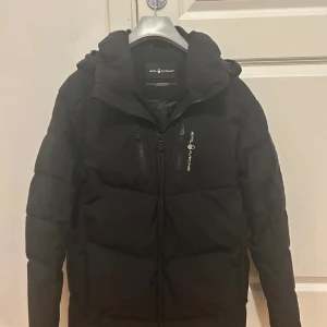 Svart pufferjacka från Sailracing - Snygg svart pufferjacka från Sailracing med huva och dragkedja. Perfekt för kyliga dagar. Jackan har en stilren design med logotypdetaljer på bröstet och ryggen. 
