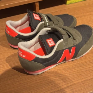 New Balance sneakers med kardborreband - Snygga New Balance sneakers i mörkblått med olivgröna detaljer och en röd logotyp. Skorna har praktiska kardborreband för enkel på- och avtagning. Perfekta för en sportig look.