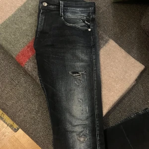 Replay jeans  - Snygga svarta jeans storlek 32/34 från Replay med slitningar och en modern look. De har en klassisk femficksdesign och knappgylf. Perfekta för en avslappnad stil med en edgy touch.