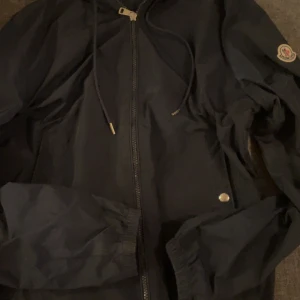 Blå vindjacka från Moncler - Snygg blå vindjacka från Moncler.Jackan har långa ärmar och en diskret logga på ärmen. Perfekt för blåsiga dagar och en stilren look.priset är inte hugget i sten!