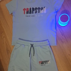 Trapstar ljusblå set - Snyggt set med TRAPSTAR ljusblå t-shirt och matchande shorts från Trapstar. T-shirten har ett tryck i rött, svart och vitt framtill. Shortsen har en elastisk midja med svart dragsko och samma tryck på benet. Perfekt för en avslappnad stil. Köp nu inför sommaren!