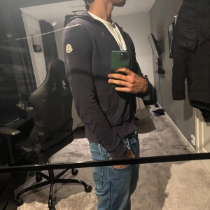 Mörkblå zip hoodie från Moncler - Säljer en stilren mörkblå hoodie från Moncler med dragkedja och huva. Tröjan har en broderad Moncler-logga på ärmen och är perfekt för en avslappnad look. Inuti tröjan står det M men passar mer som s/xs