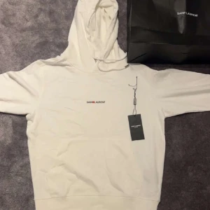 Vit hoodie från Saint Laurent - Säljer en stilren vit hoodie från Saint Laurent. Den har en klassisk design med en justerbar huva och en diskret logotyp på bröstet. Perfekt för en riktigt fet outfit! Har av er vid frågor eller andra funderingar!