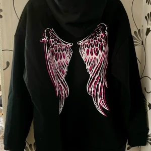 Svart hoodie med änglavingar S/M - Cool svart hoodie med texten 'I HAVE A PAIR OF WINGS' på framsidan och ett stort vingmotiv i rosa och vitt på baksidan. Perfekt för en avslappnad stil med en touch av attityd. Hoodien har en klassisk passform och en stor ficka framtill.