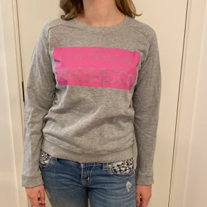 Grå sweatshirt från Black Story - Säljer en grå sweatshirt från Black Story i storlek L. Den har ett coolt rosa tryck med texten 'I'LL BE READY IN A PROSECCO'. Perfekt för en avslappnad stil med långa ärmar och mjukt material. Passar bra till jeans för en casual look.