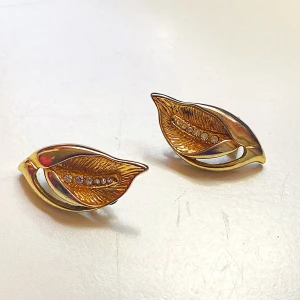 Guldiga lövformade örhängen - Säljer ett par eleganta örhängen i form av löv med en vacker guldig finish. De har en rad av gnistrande stenar längs mitten som ger en extra touch av glamour. Perfekta för att lyfta vilken outfit som helst, oavsett om det är till vardags eller fest.