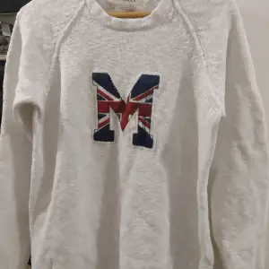 Säljer en snygg vit stickad tröja från Morris med ett stort 'M' på bröstet i Union Jack-mönster. Perfekt för höst och vinter med sin varma och bekväma design. Tröjan har långa ärmar och en klassisk passform. Passar både till vardags och festliga tillfällen!