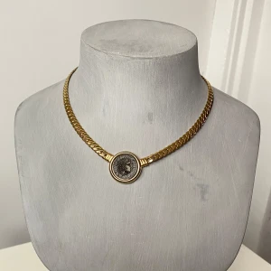 Guldhalsband med antik medaljong - Säljer ett elegant guldhalsband med en antik stil medaljong i mitten. Halsbandet har en tjock kedja och medaljongen har ett klassiskt motiv som ger en vintage känsla. Perfekt för att ge en lyxig touch till din outfit!